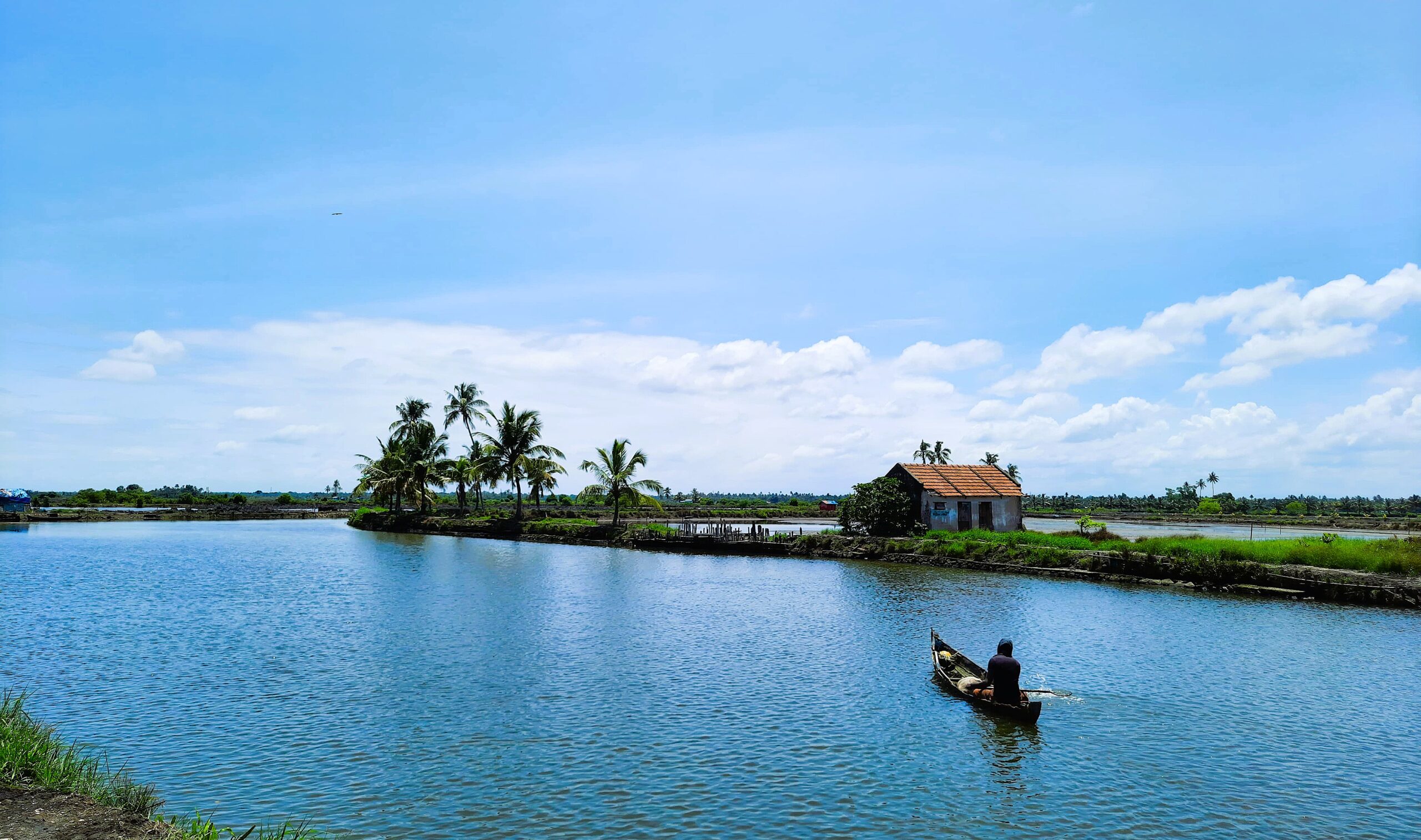 A boatman navigates the serene backwaters of Kadamakudy, with lush greenery and traditional homes lining the waterways under a clear sky.Kochi photography portfolio.